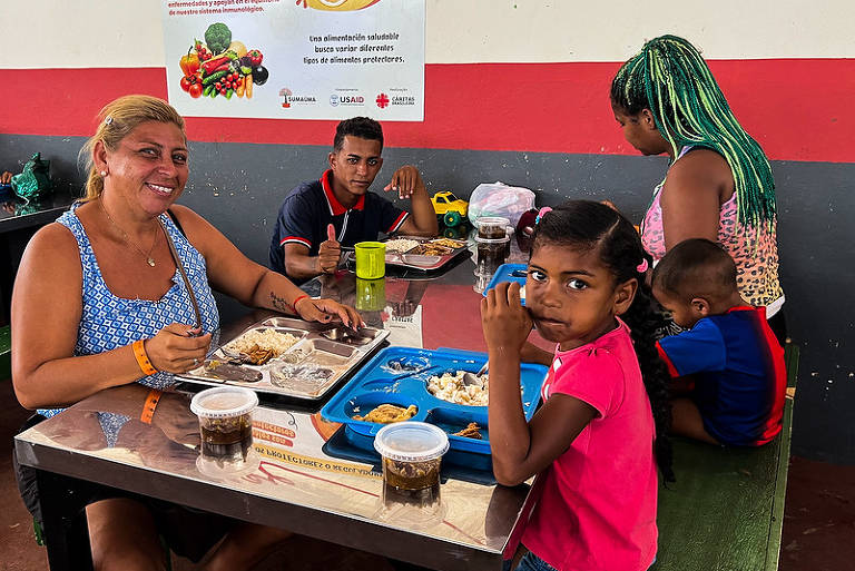 A imagem mostra um grupo de quatro pessoas sentadas em uma mesa de refeição. Uma mulher com cabelo longo e loiro está à esquerda, sorrindo e comendo. Um jovem homem está ao seu lado, olhando para a câmera. Uma menina com cabelo preso e uma blusa rosa está à frente, segurando um copo. Um menino pequeno está sentado à direita, voltado para a mesa. Ao fundo, há uma parede com uma cartaz que contém informações sobre alimentos saudáveis.