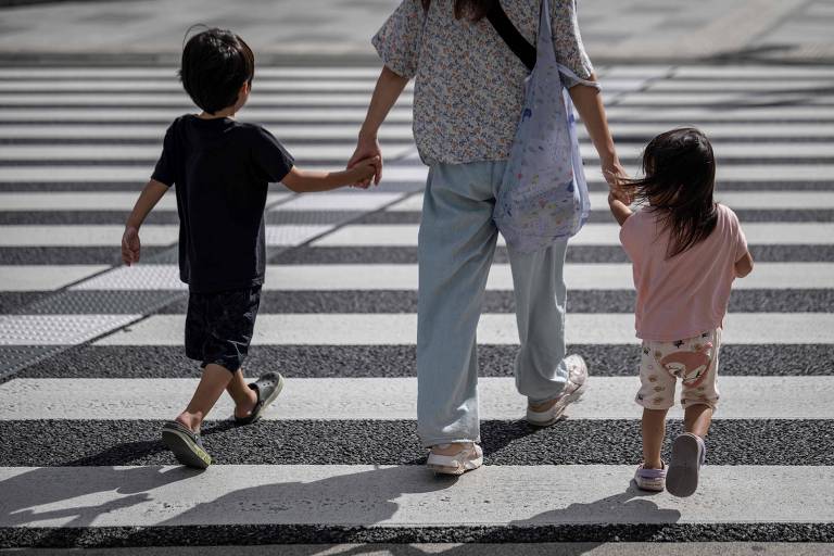 A imagem mostra uma mulher segurando as mãos de duas crianças enquanto atravessam uma faixa de pedestres. A mulher está vestindo uma blusa clara e calças largas， e carrega uma bolsa. As crianças， um menino e uma menina， estão usando roupas casuais. O menino está à esquerda， vestindo uma camiseta escura e shorts， enquanto a menina está à direita， usando uma camiseta rosa e calças claras.