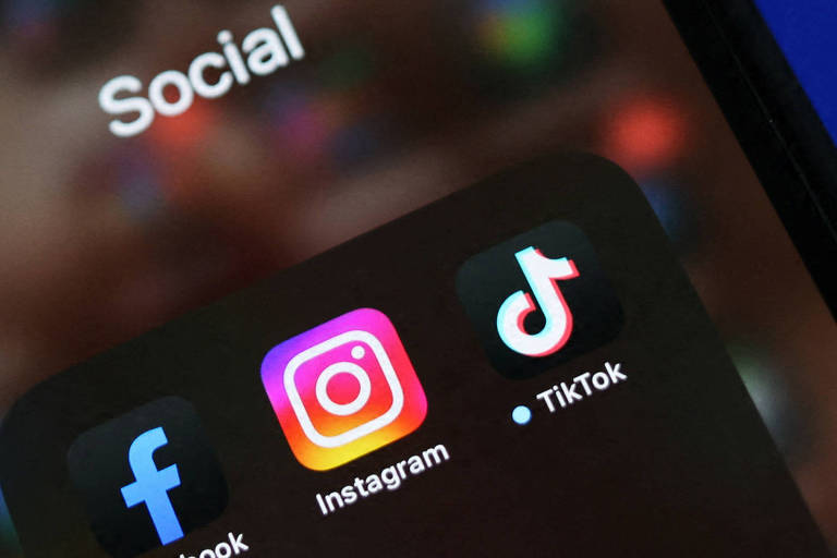 A imagem mostra um smartphone em primeiro plano， exibindo uma tela com ícones de aplicativos de redes sociais， incluindo Facebook， Instagram e TikTok. 