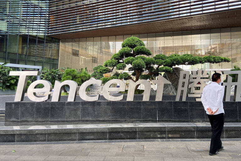 A imagem mostra uma pessoa de costas em frente ao logotipo da Tencent， que está em letras grandes e prateadas. Ao fundo， há uma planta ornamental em forma de árvore e um edifício moderno com janelas reflexivas. O ambiente é urbano e bem cuidado.