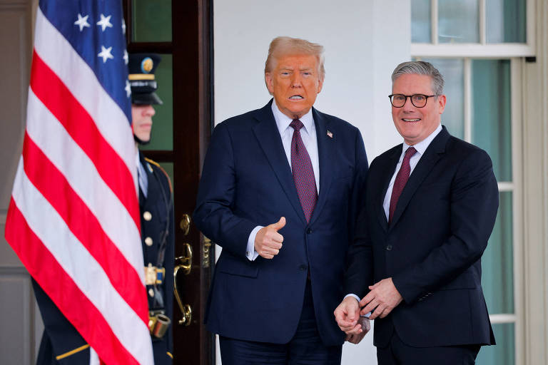 A imagem mostra duas pessoas posando do lado de fora da Casa Branca. À esquerda， há uma bandeira dos Estados Unidos. À direita， um homem de terno escuro e gravata clara está sorrindo e segurando a mão do outro homem， que está vestido com um terno escuro e gravata vermelha. O homem à direita faz um gesto de positivo com o polegar.