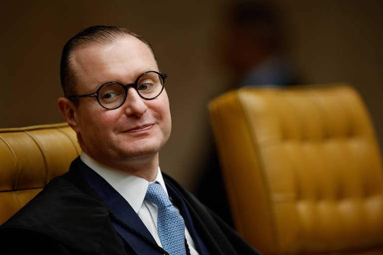 Um homem com cabelo curto e liso， usando óculos redondos e um terno escuro， está sentado em uma cadeira de couro marrom. Ele tem uma expressão serena e está olhando para a frente. Ao fundo， há uma leve desfocagem que sugere um ambiente formal.