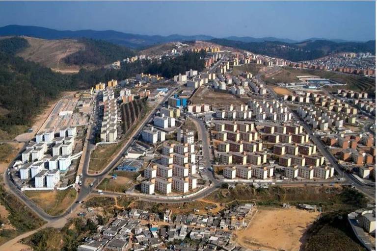 A imagem mostra uma vista aérea de um grande conjunto habitacional, com prédios de vários andares dispostos em áreas organizadas. À esquerda, há uma área com prédios de cores claras e, à direita, uma área com prédios em tons mais variados. Ao fundo, é possível ver colinas e uma área urbana mais densa. A parte inferior da imagem apresenta uma área com construções mais simples, possivelmente uma favela.