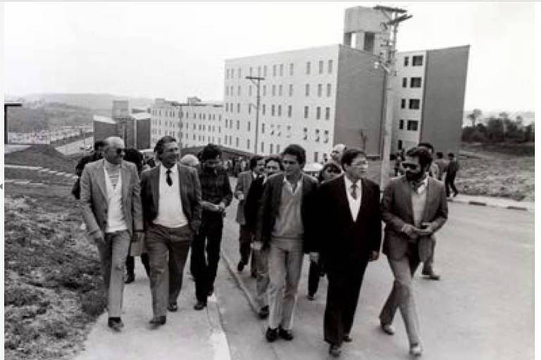 Um grupo de homens caminha em uma rua, com prédios ao fundo. Eles estão vestidos de maneira formal e casual, alguns usando paletós e gravatas. O ambiente parece ser urbano, com uma construção de prédios de vários andares visível. O céu está nublado, e a cena transmite um ar de atividade e interação entre os indivíduos.