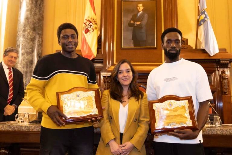 A imagem mostra uma cerimônia de premiação em um ambiente formal. Duas pessoas， um homem à esquerda com um suéter preto e amarelo e outro à direita com uma camiseta branca， estão segurando placas de reconhecimento. Entre eles， uma mulher com um blazer amarelo sorri. Ao fundo， há um retrato emoldurado de um homem e bandeiras. Um homem mais velho， vestido de terno， observa ao fundo.