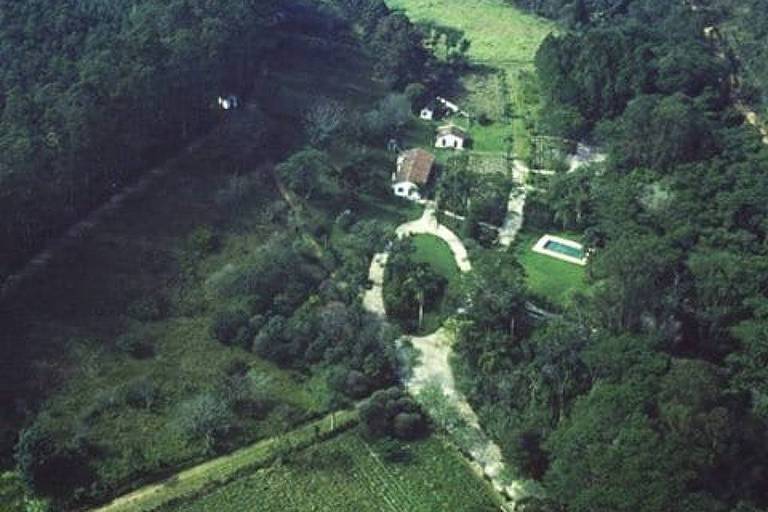 Imagem aérea de uma propriedade rural cercada por vegetação densa. No centro, há uma casa com telhado vermelho e uma piscina ao lado. Caminhos de terra se entrelaçam na área, levando a diferentes partes da propriedade. A vegetação ao redor é predominantemente verde, com árvores e arbustos.