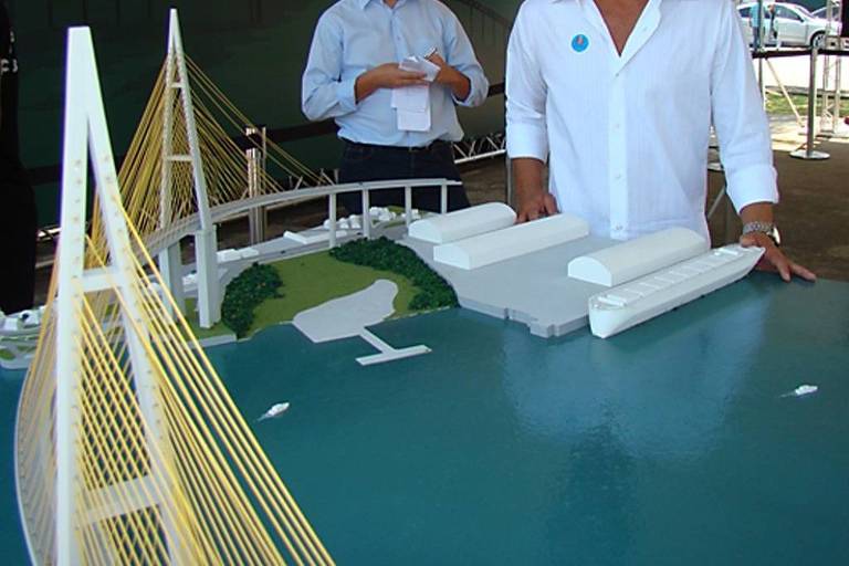 A imagem mostra uma maquete de uma ponte com cabos amarelos e uma estrutura portuária ao lado. Um homem em uma camisa branca está em pé ao lado da maquete， enquanto outras pessoas estão ao fundo， algumas segurando papéis. A maquete é apresentada sobre uma mesa， com um fundo que parece ser um evento ao ar livre.