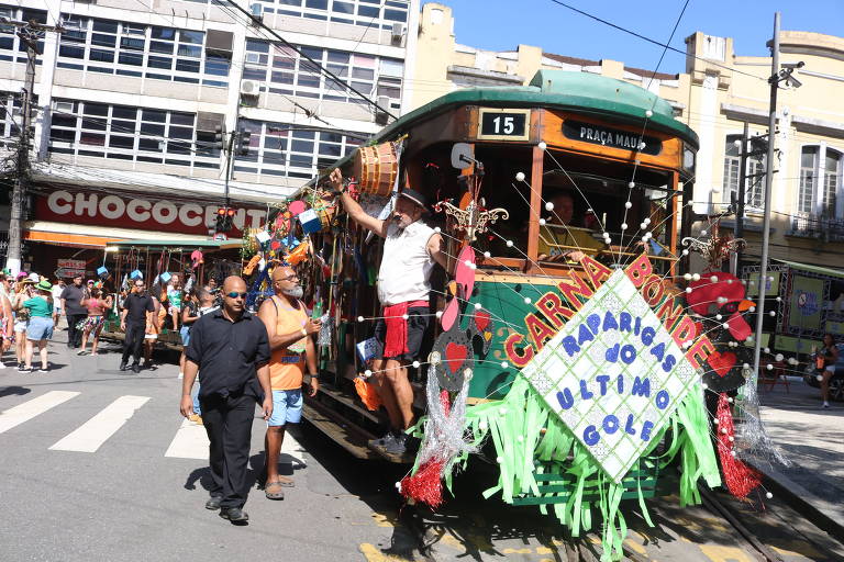 A imagem mostra um bonde decorado para o carnaval， com várias fitas e enfeites coloridos. Um homem está em pé na parte externa do bonde， enquanto outro homem caminha ao lado. Ao fundo， há uma loja chamada 039;Chococita039; e várias pessoas participando da festa na rua. O bonde exibe a placa 039;15039; e a frase 039;CARNAVAL BONDE - PARABÉNS AO ÚLTIMO GOL039;.