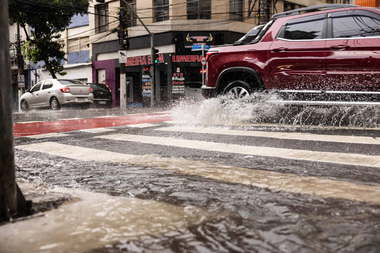 A imagem mostra uma rua urbana inundada， com água acumulada sobre a faixa de pedestres. Um carro vermelho está passando， levantando respingos de água. Ao fundo， há prédios e outros veículos， além de uma árvore na lateral esquerda.