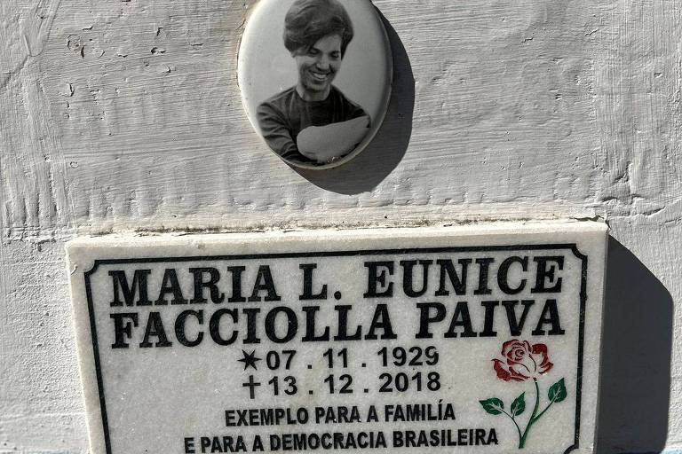 A imagem mostra uma placa em homenagem a Maria L. Eunice Facciolla Paiva， com a data de nascimento 07.11.1929 e a data de falecimento 13.12.2018. Acima da placa， haacute; uma foto em preto e branco de uma mulher segurando um livro. A placa conteacute;m a frase 039;Exemplo para a famiacute;lia e para a democracia brasileira039; e um desenho de uma rosa.