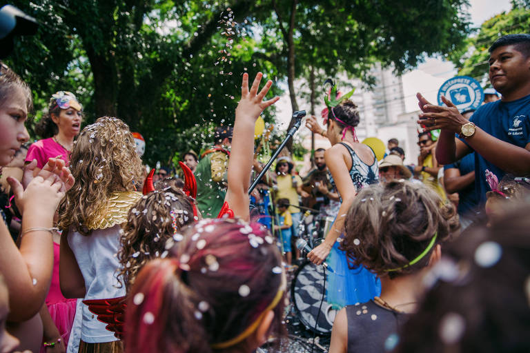 A imagem mostra um grupo de crianças e adultos em uma festa de Carnaval ao ar livre. Algumas crianças estão vestidas com fantasias coloridas e acessórios brilhantes， enquanto outras estão aplaudindo e se divertindo. Há confetes caindo no ar e um microfone em um suporte， indicando que há uma apresentação musical. Ao fundo， árvores e pessoas assistindo à festa.