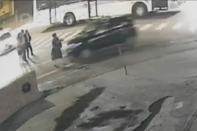 A imagem mostra uma cena de um acidente de trânsito em uma rua. Um carro preto está em movimento， aparentemente em alta velocidade， enquanto um grupo de pessoas está parado na calçada. Ao fundo， um ônibus está estacionado. A imagem parece ter sido capturada por uma câmera de segurança， e há uma névoa ou fumaça visível na cena.