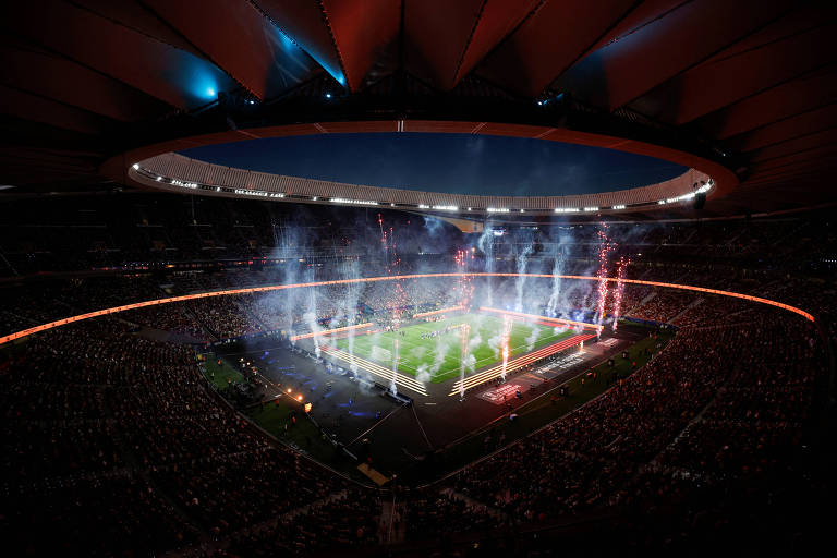 Imagem de um estádio iluminado durante um evento noturno， com fogos de artifício e fumaça no ar. O campo de futebol está visível， cercado por uma grande quantidade de espectadores nas arquibancadas. O teto do estádio é visível， com iluminação azul e detalhes arquitetônicos.