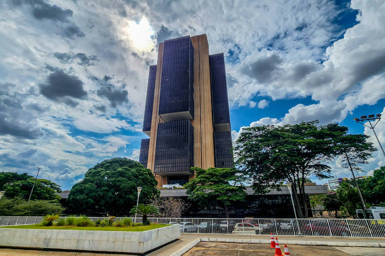 A imagem mostra um edifício moderno de grande altura, com uma fachada predominantemente escura e detalhes em madeira. O céu está nublado, com nuvens brancas e cinzas, e há uma leve iluminação solar. Na parte inferior, há árvores verdes e uma área pavimentada com algumas barreiras de segurança visíveis.