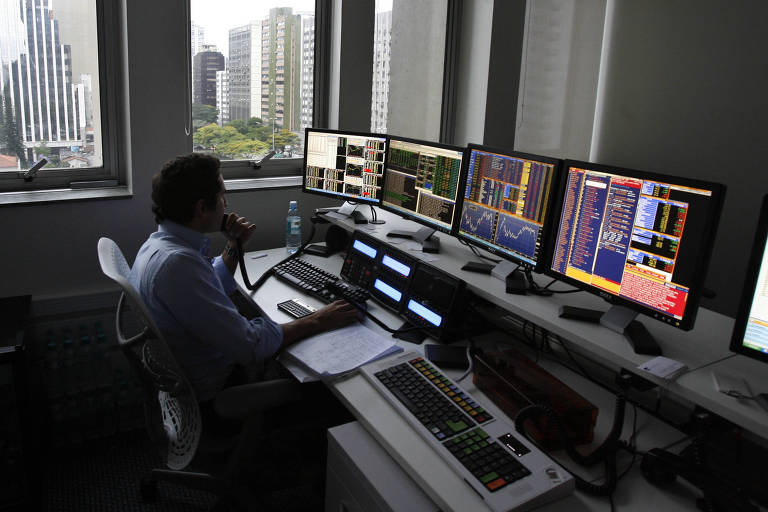 A imagem mostra um ambiente de trabalho em um escritório financeiro. Um homem está sentado em frente a várias telas de computador que exibem gráficos e dados financeiros. Ele parece concentrado， com uma mão segurando o telefone e a outra， uma folha de papel. Ao fundo， há grandes janelas que oferecem uma vista da cidade， com prédios altos e vegetação visível