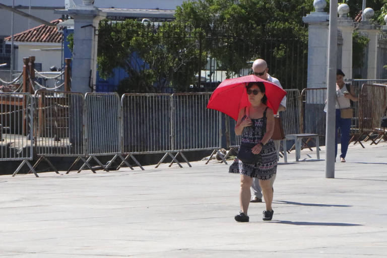 Uma mulher caminha em um calçadão， segurando um guarda-sol vermelho. Ela usa um vestido estampado e sandálias. Ao fundo， um homem com cabelo grisalho caminha atrás dela. Há cercas de metal ao redor e algumas árvores visíveis.