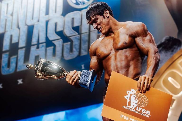 Um competidor de fisiculturismo está em um palco segurando um troféu e um cartão. Ele está em pose de vitória， exibindo um corpo musculoso. Ao fundo， há uma tela com o nome 039;ARNOLD CLASSIC039; e o logotipo da IFBB.