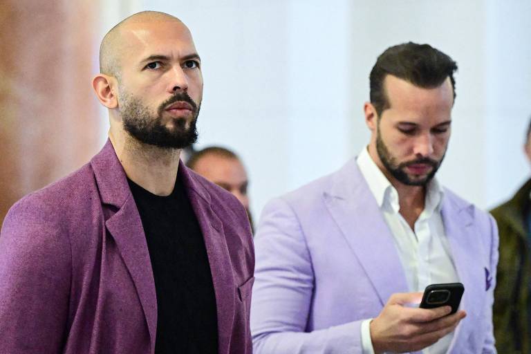 A imagem mostra dois homens em trajes formais. O homem à esquerda tem cabelo raspado， barba e usa um paletó roxo sobre uma camiseta preta. O homem à direita tem cabelo escuro e usa um paletó lilás sobre uma camisa branca， enquanto olha para um celular. Ao fundo， há outras pessoas desfocadas.