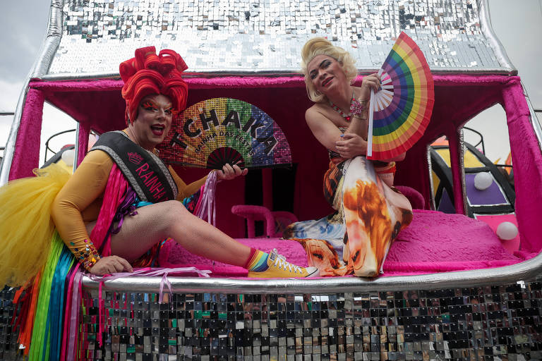 Duas drag queens estão sentadas em um carro alegórico decorado com espelhos e cores vibrantes. A primeira drag， à esquerda， usa um vestido colorido com uma saia volumosa e uma peruca vermelha. Ela segura um leque colorido e exibe um sorriso. A segunda drag， à direita， veste um vestido laranja e segura um leque com as cores do arco-íris. Ambas estão em um ambiente festivo， com detalhes brilhantes ao fundo.