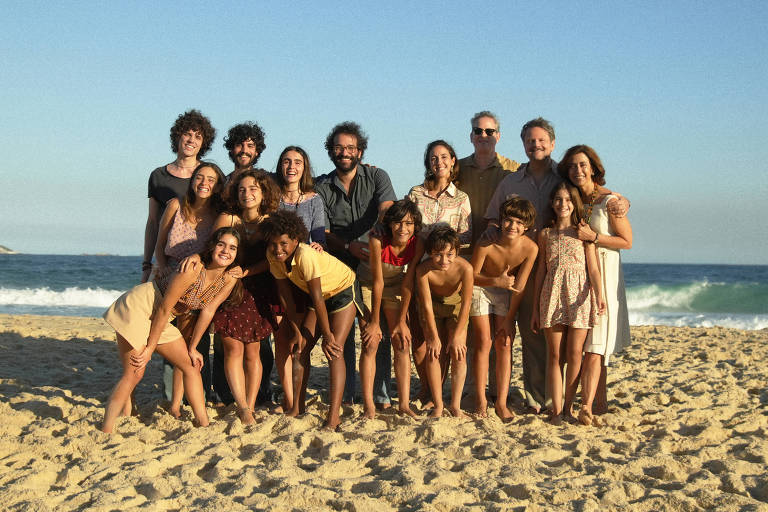 Uma foto de um grupo de 17 pessoas posando na praia. Elas estão em pé e agachadas na areia， com o mar ao fundo. A maioria das pessoas está sorrindo e vestindo roupas leves de verão. O céu está claro e há ondas no mar.