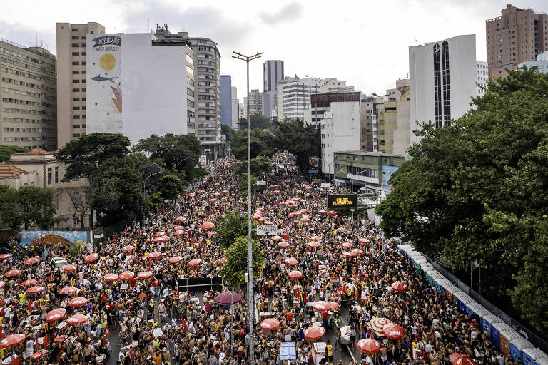 A imagem mostra uma grande multidão reunida em uma área urbana， com prédios altos ao fundo. As pessoas estão em uma rua， muitas delas sob guarda-sóis vermelhos. Árvores estão visíveis ao lado da rua， e há uma atmosfera de agitação e atividade. O céu está nublado， sugerindo um dia com possibilidade de chuva.