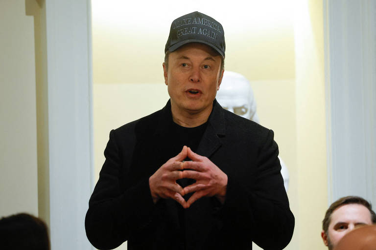 Musk está em pé， falando em um evento. Ele usa um casaco preto e um boné cinza com a frase 039;Make America Great Again039;. Ao fundo， há uma escultura branca e outras pessoas parcialmente visíveis.