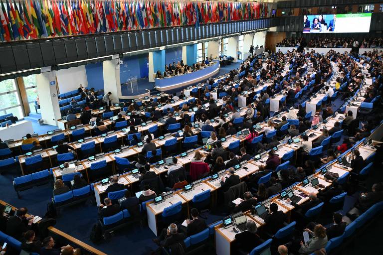 A imagem mostra uma grande sala de conferências com várias pessoas sentadas em mesas dispostas em fileiras. No fundo， há uma parede decorada com bandeiras de diferentes países. Um painel de apresentação é visível à direita， e a iluminação é natural， proveniente de janelas amplas.