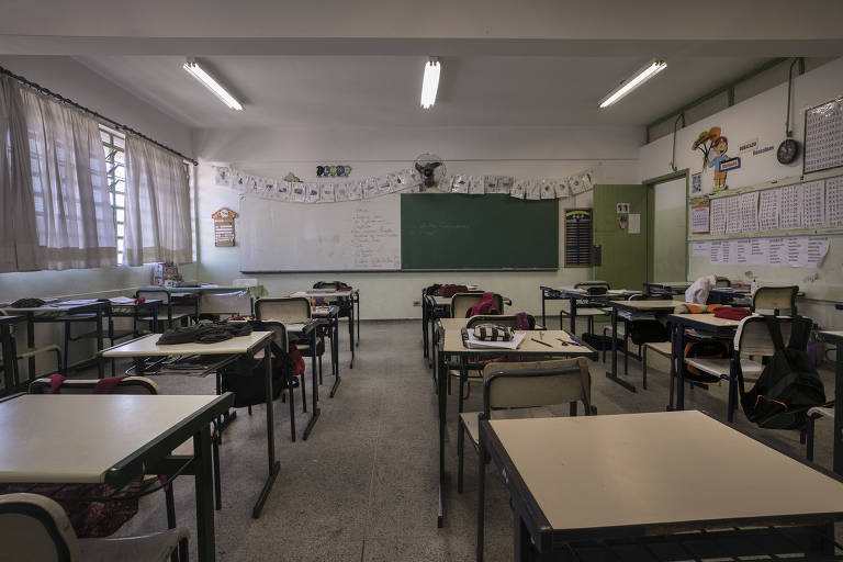 A imagem mostra uma sala de aula vazia com mesas e cadeiras dispostas em fileiras. Há uma lousa verde na parede frontal， com algumas anotações visíveis. As janelas têm cortinas claras e há uma decoração na parte superior da sala. O ambiente é bem iluminado por lâmpadas fluorescentes no teto.