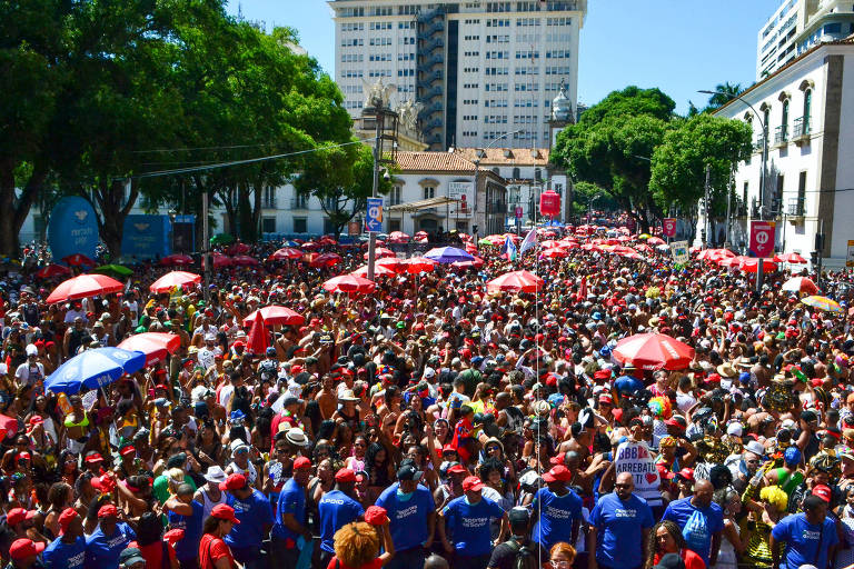 A imagem mostra uma grande multidão reunida em um evento ao ar livre， com pessoas usando camisetas azuis e chapéus vermelhos. Há muitos guarda-sóis vermelhos espalhados pela área， e edifícios altos ao fundo. A cena é vibrante e cheia de energia， com árvores verdes ao redor.