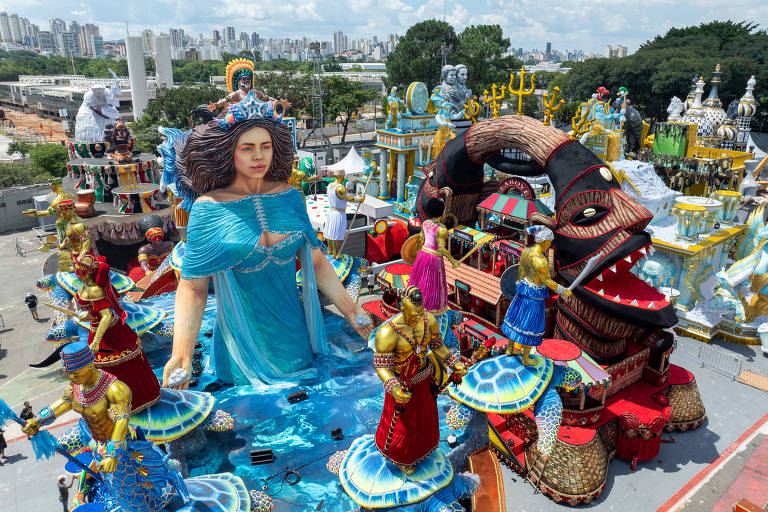 A imagem mostra um grande carro alegórico de carnaval com uma figura central feminina vestida de azul， representando uma deusa do mar. Ao redor， há várias estátuas e figuras em trajes coloridos， algumas em posições de dança， e outras em trajes tradicionais. O cenário é vibrante， com elementos decorativos que incluem peixes， conchas e uma grande boca de um dragão. Ao fundo， a cidade é visível com prédios e árvores.