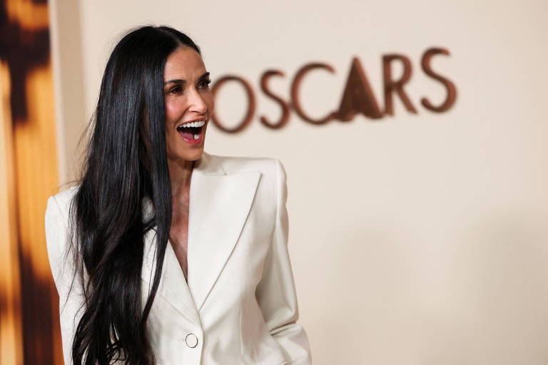 Uma mulher com cabelo longo e liso， vestindo um blazer branco， está sorrindo e se divertindo em um evento. Ao fundo， há a palavra 039;OSCARS039; em letras grandes e estilizadas.