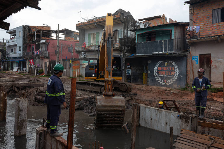 A imagem mostra uma obra de construção em uma área urbana. No primeiro plano， há um trabalhador em pé em uma estrutura de concreto， observando uma escavadeira amarela que está prestes a levantar um pedaço de material. Ao fundo， vê-se uma série de casas de diferentes estilos， algumas com varandas e outras com paredes de tijolos expostos. O céu está nublado， sugerindo um clima chuvoso.