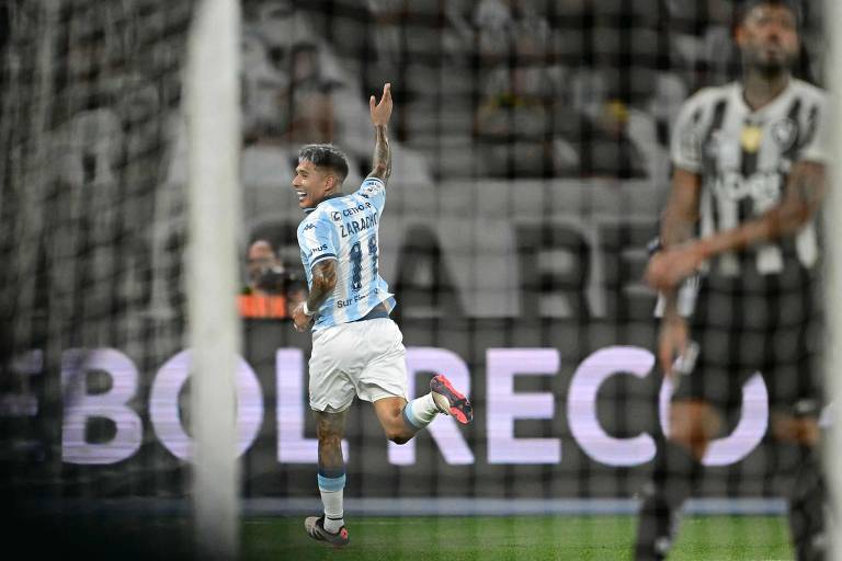 Um jogador de futebol， vestindo uma camisa azul e branca com o número 11， está comemorando um gol. Ele está correndo em direção à torcida， levantando o braço em sinal de celebração. Ao fundo， é possível ver jogadores adversários com camisas listradas em preto e branco. O ambiente é de um estádio de futebol， com uma iluminação intensa e uma tela exibindo a palavra 039;BOLERCO039;.