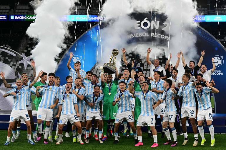 Um grupo de jogadores de futebol， vestindo uniformes listrados em azul e branco， celebra a conquista de um troféu. Eles estão em um campo de futebol， com alguns jogadores levantando o troféu acima de suas cabeças. Ao fundo， há fumaça branca e uma grande estrutura com o nome 039;Recopa039;. A equipe parece estar muito animada e feliz.