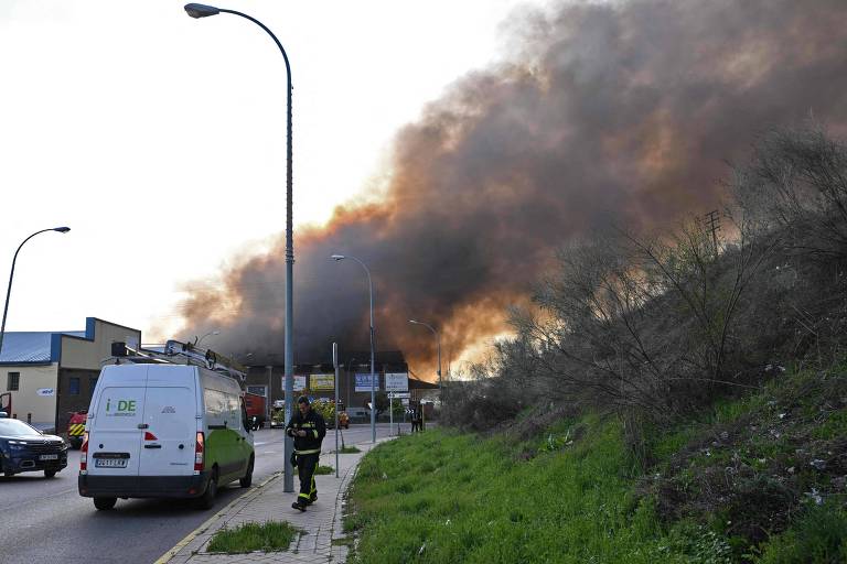 A imagem mostra uma cena de incêndio em uma área industrial. Grandes nuvens de fumaça preta se elevam no céu, indicando um incêndio significativo. No primeiro plano, há um caminhão de bombeiros e um veículo branco estacionado na calçada. Um bombeiro está visível, caminhando em direção ao local do incêndio. O ambiente é urbano, com vegetação ao lado da estrada.