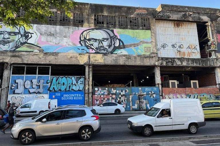 A imagem mostra a fachada de um edifício em um ambiente urbano. A parede é decorada com grafites coloridos， incluindo rostos e frases. Há veículos estacionados na rua em frente ao edifício， incluindo um carro branco e uma van. O céu está claro e há árvores na parte superior da imagem.