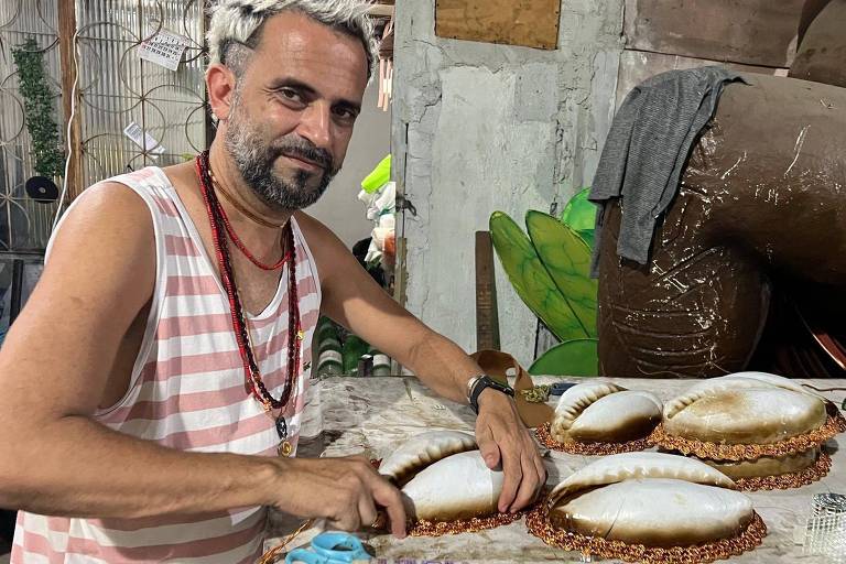 Um homem com cabelo grisalho e barba， vestindo uma camiseta listrada， está sentado à mesa， trabalhando em conchas decorativas. Ele está concentrado， manipulando as conchas que estão dispostas sobre a mesa. Ao fundo， há uma parede com elementos decorativos e uma planta verde visível.