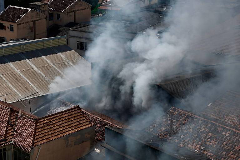 A imagem mostra uma vista aérea de um incêndio em uma área urbana， com fumaça densa saindo de um edifício. O telhado de cerâmica de várias casas é visível， e a fumaça se espalha pelo ar， obscurecendo parcialmente a visão. O céu está claro， contrastando com a fumaça.