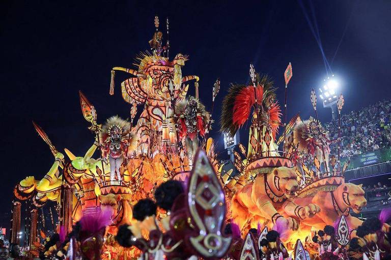 A imagem mostra um grande carro alegórico iluminado， decorado com figuras de leões e elementos coloridos， durante um desfile de Carnaval à noite. O fundo apresenta uma multidão assistindo ao evento， com luzes brilhantes e uma atmosfera festiva.