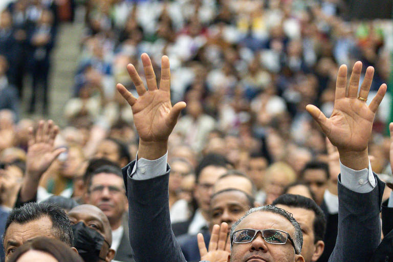 A imagem mostra uma grande audiência em um evento， com várias pessoas levantando as mãos. No primeiro plano， há um homem de óculos e uma mulher com cabelo longo e escuro. Ao fundo， é possível ver uma multidão de pessoas， algumas também levantando as mãos， em um ambiente interno com cadeiras dispostas em fileiras.