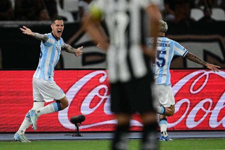 Na imagem， um jogador com camisa listrada em azul e branco está comemorando um gol， com os braços abertos e expressando alegria. Ao fundo， outro jogador com a camisa número 5 também está presente. A cena ocorre em um estádio， com um painel publicitário da Coca-Cola visível.