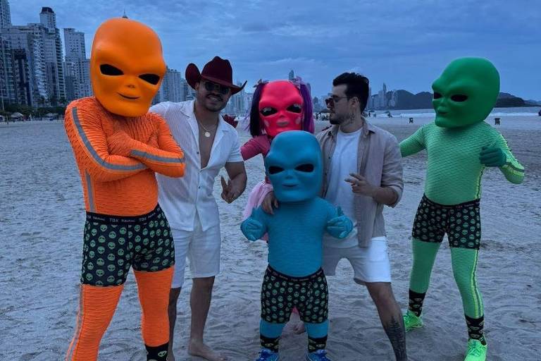 Cinco pessoas estão na praia durante o crepúsculo. Quatro delas estão vestidas com trajes coloridos de alienígenas， em tons de laranja， verde， azul e rosa， enquanto uma pessoa está vestindo um traje branco e chapéu. O fundo mostra um céu nublado e a silhueta de prédios ao longe.