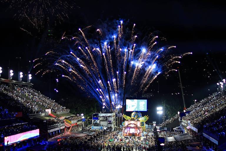 A imagem mostra um grande evento noturno com uma multidão reunida em um espaço aberto. Fogos de artifício iluminam o céu， criando um espetáculo colorido. No fundo， há uma grande tela de vídeo e estruturas de palco. As arquibancadas estão cheias de pessoas， e as luzes do evento brilham intensamente.