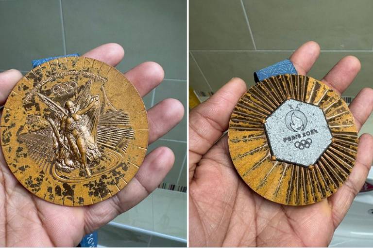 A imagem mostra uma medalha olímpica sendo segurada por uma mão. O lado frontal da medalha apresenta uma figura esculpida de um atleta， com detalhes que incluem os anéis olímpicos e um fundo com linhas radiantes. O verso da medalha é hexagonal， com um design em relevo que inclui a inscrição 039;PARIS 2024039; e os anéis olímpicos. A medalha é de cor dourada e possui uma fita azul.