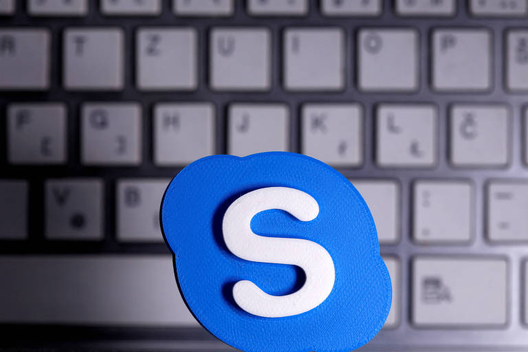 A imagem mostra o logotipo do Skype， que é um ícone azul com a letra 039;S039; branca， em primeiro plano. Ao fundo， há um teclado de computador com teclas visíveis em tons de cinza.