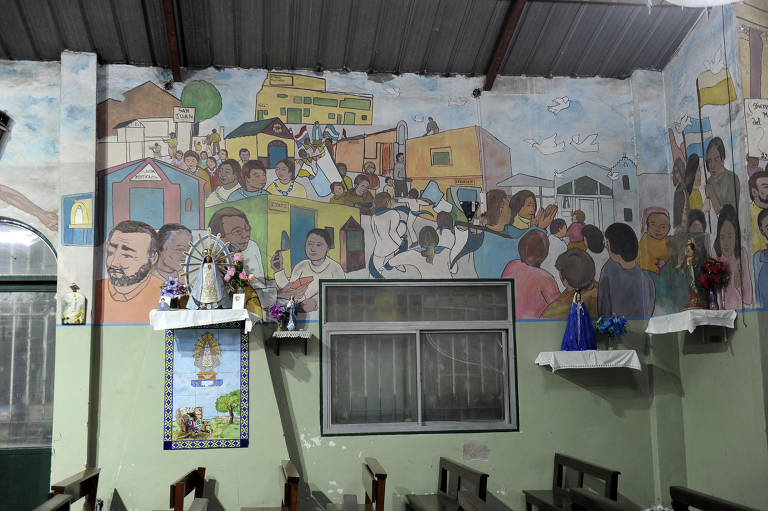 A imagem mostra um mural colorido em uma igreja, retratando uma cena comunitária com várias pessoas, casas e árvores. O mural ocupa a parede e apresenta figuras humanas em diferentes posturas, algumas com expressões alegres. Na parte inferior, há uma mesa com objetos decorativos e flores, e uma imagem religiosa emoldurada. A iluminação é suave, e o teto é de metal.