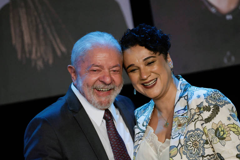 A imagem mostra um homem e uma mulher sorrindo em um evento. O homem tem cabelo grisalho e usa um terno escuro com uma gravata. A mulher tem cabelo curto e escuro， e está vestindo uma jaqueta com estampas florais. Ambos parecem felizes e estão próximos um do outro， com um fundo desfocado que sugere um ambiente de evento.