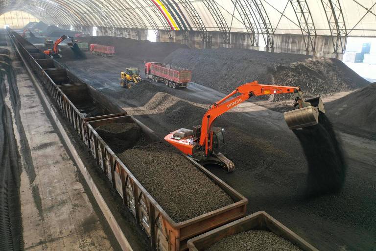 A imagem mostra um armazém coberto onde estão armazenados grandes montes de material， possivelmente minério ou carvão. Vários caminhões e equipamentos pesados， como escavadeiras， estão em operação， carregando o material em vagões de trem. O ambiente é amplo， com uma estrutura de cobertura que permite a entrada de luz.