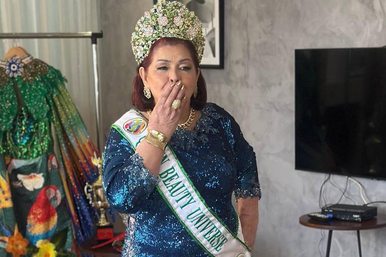 Uma mulher está em pé em um ambiente interno, usando um vestido azul brilhante e uma coroa. Ela está fazendo um gesto de beijo com a mão e tem uma faixa que diz 'Miss'. Ao fundo, há uma decoração colorida e uma televisão na parede.