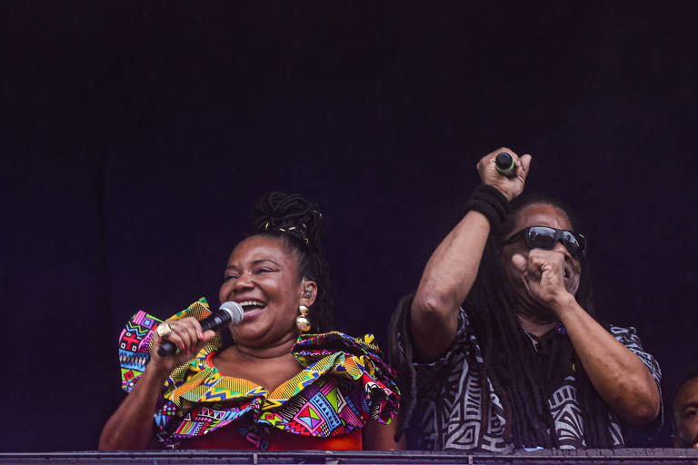 Duas pessoas estão se apresentando em um palco. A mulher à esquerda está usando uma blusa colorida com estampas africanas e segurando um microfone, sorrindo. O homem à direita está usando óculos escuros e uma camiseta preta com padrões, levantando a mão em um gesto de celebração. O fundo é escuro, sugerindo um ambiente de show.